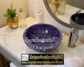 May include: A purple amethyst stone sink bowl with a rough, textured exterior. The bowl is sitting on a white countertop in a bathroom setting. The text "UniqueHandicraftsIND" is visible on the bowl.