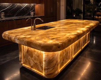 May include: A kitchen island with a polished, golden countertop. The island has a built-in cooktop and faucet. The base is illuminated with warm lighting, creating a sophisticated atmosphere. The island is made of a marble-like material. The countertop is approximately 200 cm long.