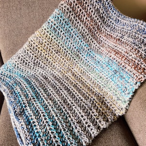 May include: A handmade crochet blanket in shades of blue, gray, brown, and white. The blanket is folded over a brown upholstered chair. The yarn has a textured appearance, with visible stitches creating a striped pattern. The blanket is a cozy home decor item.