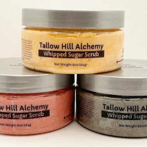 May include: Three jars of whipped sugar scrub with silver lids. The jars are labeled "Tallow Hill Alchemy Whipped Sugar Scrub" and have a net weight of 8oz/224g. The scrubs are in the colors orange, pink, and gray.