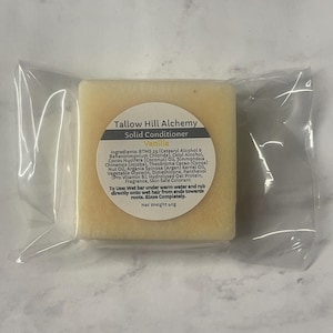 Op de afbeelding: Een witte staaf vaste conditioner met het label "Tallow Hill Alchemy Solid Conditioner Vanilla" en een lijst met ingrediënten. De staaf is verpakt in een doorzichtige plastic zak.