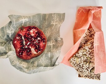 May include: Two pieces of food wrapped in reusable food wraps. A half of a pomegranate is wrapped in a gray wrap with a white tree pattern. A piece of granola is wrapped in a pink wrap with a white tree pattern.