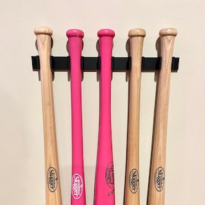 May include: Five wooden baseball bats with pink and brown paint. The bats are hanging on a black wall mount. The bats have the Louisville Slugger logo and the text "Louisville Slugger Museum & Factory". One bat has the text "Reds vs Cardinals June 8th, 2013".