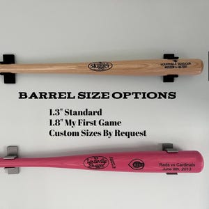 May include: Two wooden baseball bats mounted on a white wall. The top bat is natural wood with a 1.3 inch barrel. The bottom bat is pink with a 1.8 inch barrel. The text "BARREL SIZE OPTIONS" is printed below the bats. The text "1.3" Standard, 1.8" My First Game, Custom Sizes By Request" is printed below the title. The text "Louisville Slugger" is printed on both bats.