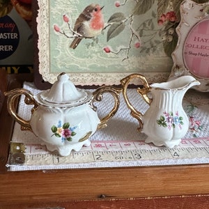 May include: A white porcelain tea set with gold trim, including a lidded sugar bowl and a creamer. Both pieces are decorated with small floral bouquets in pink, blue, and yellow. The set is placed on a white surface with a measuring tape.