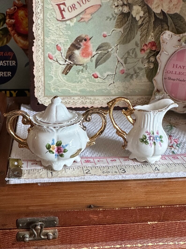 May include: A white porcelain tea set with gold trim, including a lidded sugar bowl and a creamer. Both pieces are decorated with small floral bouquets in pink, blue, and yellow. The set is placed on a white surface with a measuring tape.