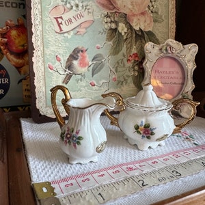 May include: A white porcelain creamer and sugar bowl set with gold trim and floral accents. The set is displayed on a white cloth with a measuring tape. A decorative picture frame and a floral print background are also visible.