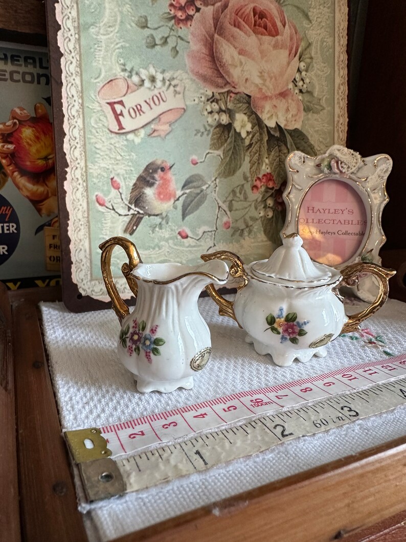 May include: A white porcelain creamer and sugar bowl set with gold trim and floral accents. The set is displayed on a white cloth with a measuring tape. A decorative picture frame and a floral print background are also visible.