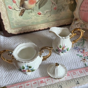 May include: A white porcelain sugar bowl and creamer set with gold trim and floral accents. The sugar bowl has a lid and is open, next to a creamer with a handle. A measuring tape shows inches and centimeters.