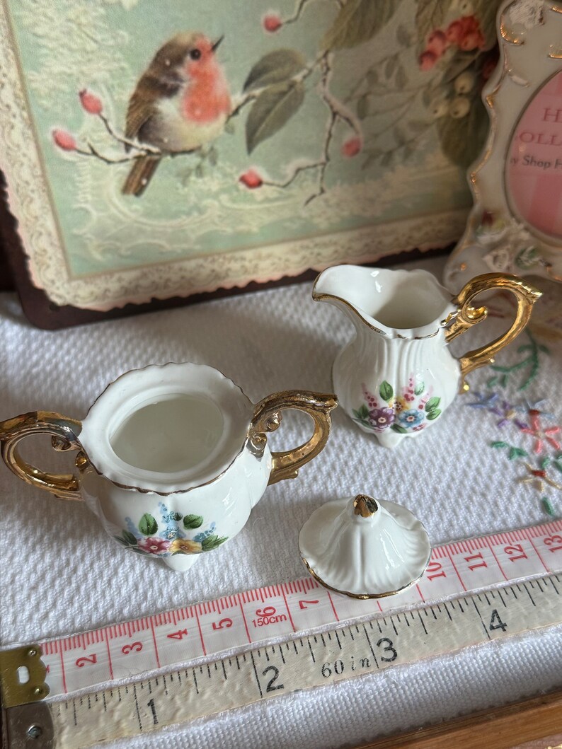 May include: A white porcelain sugar bowl and creamer set with gold trim and floral accents. The sugar bowl has a lid and is open, next to a creamer with a handle. A measuring tape shows inches and centimeters.