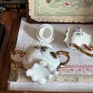 May include: A white porcelain sugar bowl with gold handles and floral accents, next to a matching lid and a bird-shaped creamer. The sugar bowl is approximately 2 inches wide. The set is on a white cloth.
