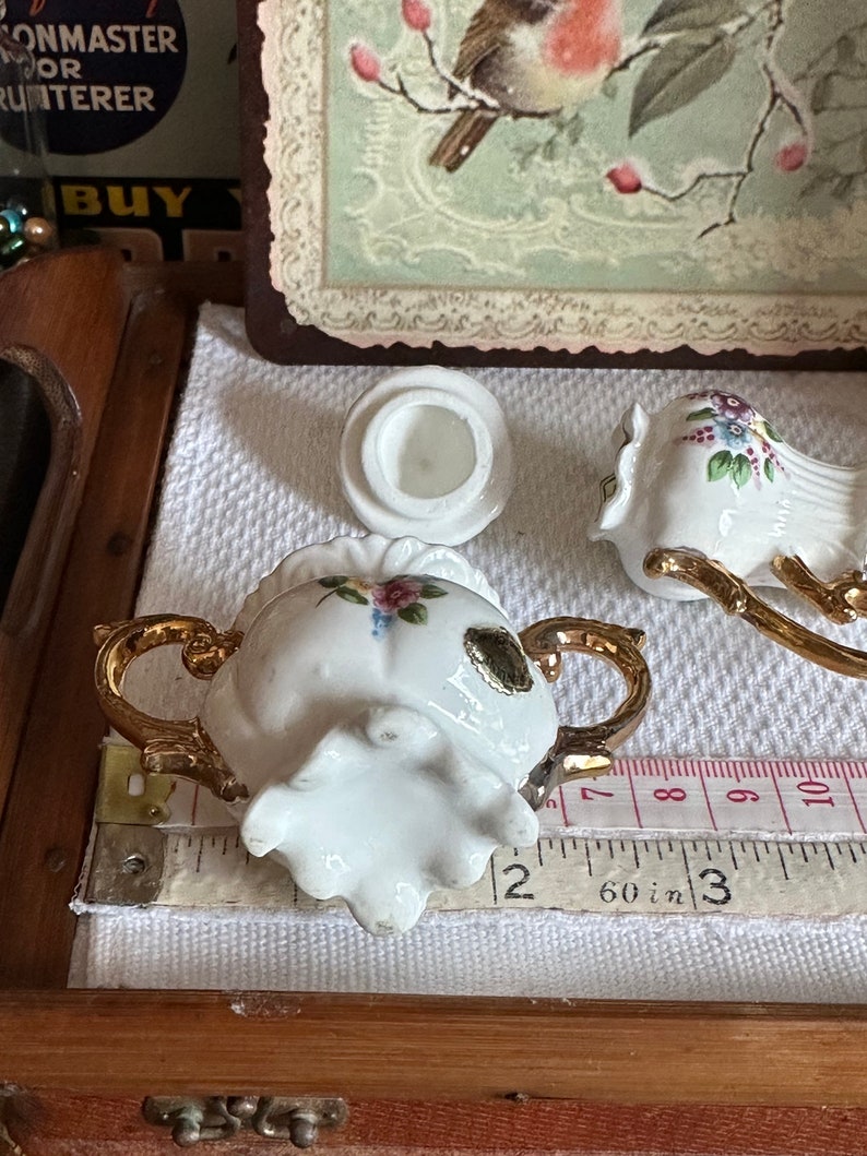 May include: A white porcelain sugar bowl with gold handles and floral accents, next to a matching lid and a bird-shaped creamer. The sugar bowl is approximately 2 inches wide. The set is on a white cloth.