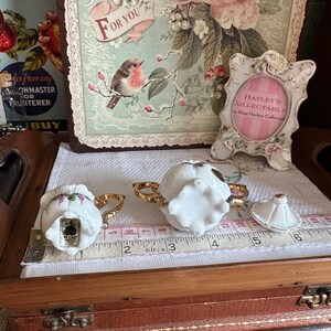 May include: Two small, white porcelain teacups with gold handles and floral accents sit on a white cloth. A matching lid is nearby. A measuring tape shows inches. A decorative picture frame and a floral print are in the background.