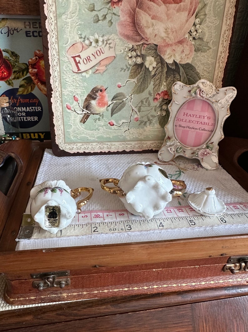 May include: Two small, white porcelain teacups with gold handles and floral accents sit on a white cloth. A matching lid is nearby. A measuring tape shows inches. A decorative picture frame and a floral print are in the background.