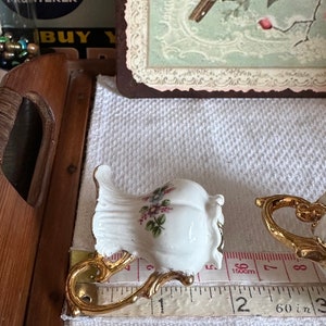 May include: White porcelain teacup with gold-colored handle and floral design. The teacup is approximately 2 inches tall. A measuring tape is placed next to the teacup.