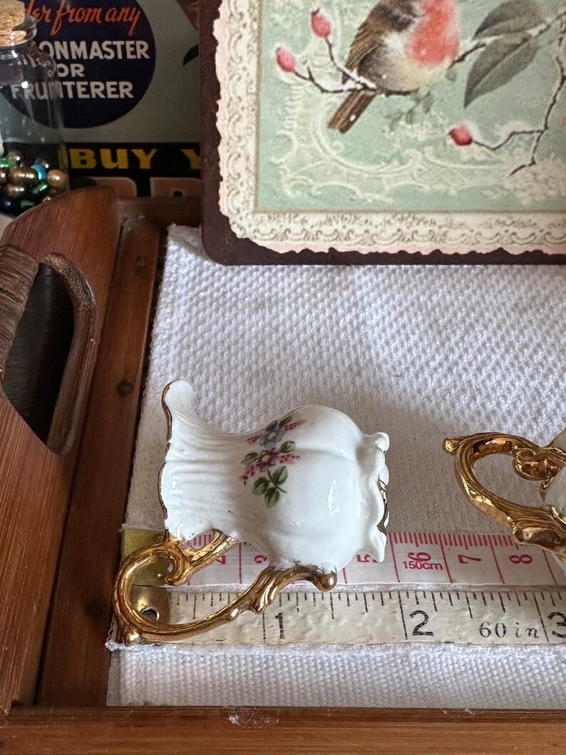 May include: White porcelain teacup with gold-colored handle and floral design. The teacup is approximately 2 inches tall. A measuring tape is placed next to the teacup.
