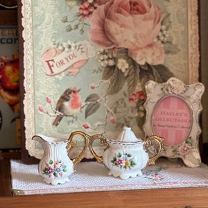May include: A white porcelain creamer and sugar bowl set with gold handles and floral accents. The set is displayed on a white cloth with a floral pattern, against a backdrop of a vintage floral print.
