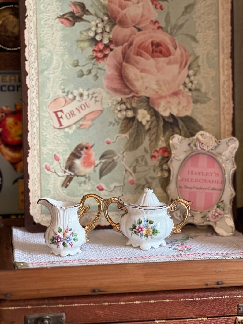 May include: A white porcelain creamer and sugar bowl set with gold handles and floral accents. The set is displayed on a white cloth with a floral pattern, against a backdrop of a vintage floral print.