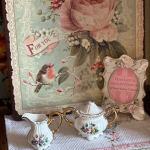 May include: A white porcelain creamer and sugar bowl set with gold handles and floral accents. The set is displayed on a white cloth with a floral design. A decorative picture with a bird and the words "For You" is in the background.