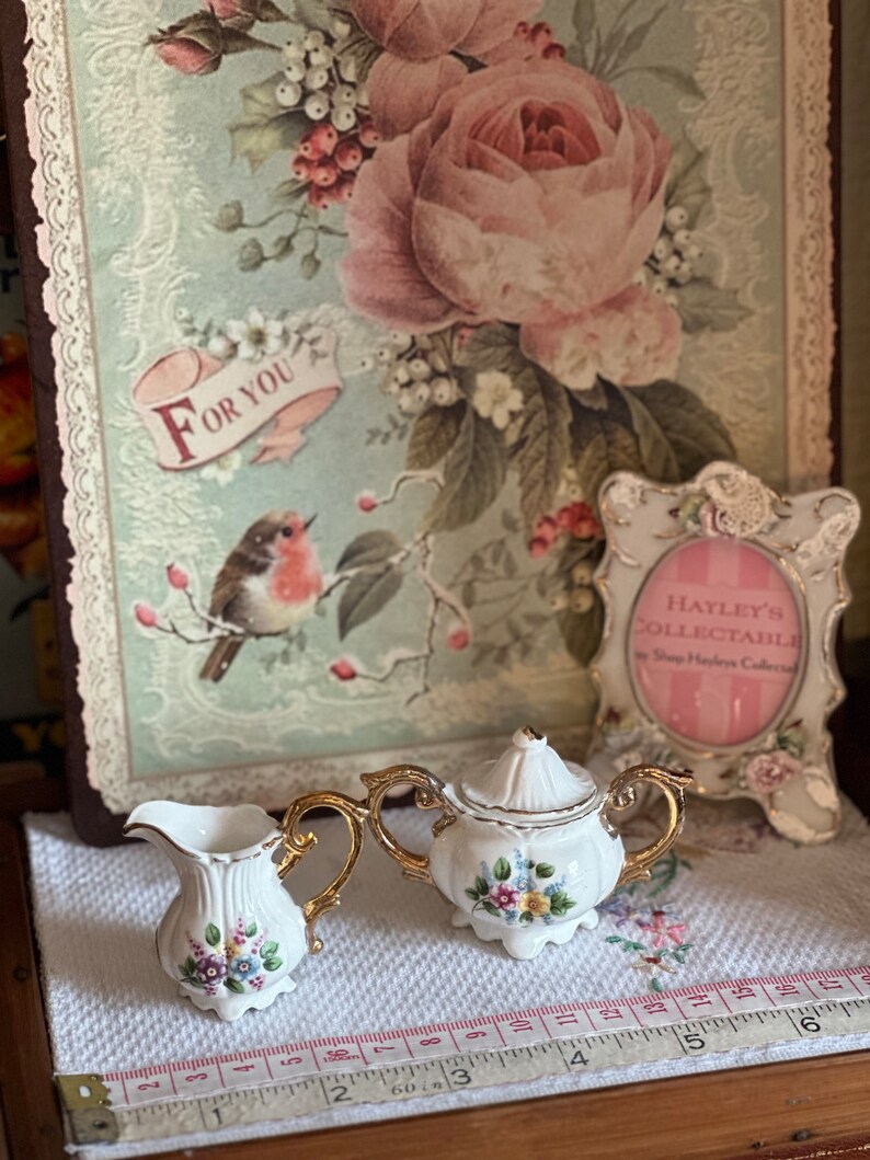 May include: A white porcelain creamer and sugar bowl set with gold handles and floral accents. The set is displayed on a white cloth with a floral design. A decorative picture with a bird and the words "For You" is in the background.
