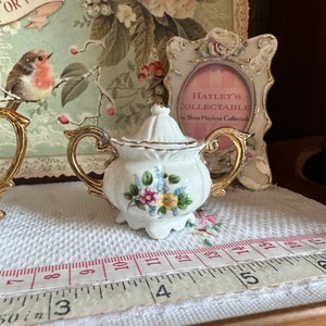 May include: A small, white porcelain sugar bowl with a lid, gold trim, and floral accents. The sugar bowl is approximately 3 inches wide. The background includes a decorative frame and a vintage-style print.