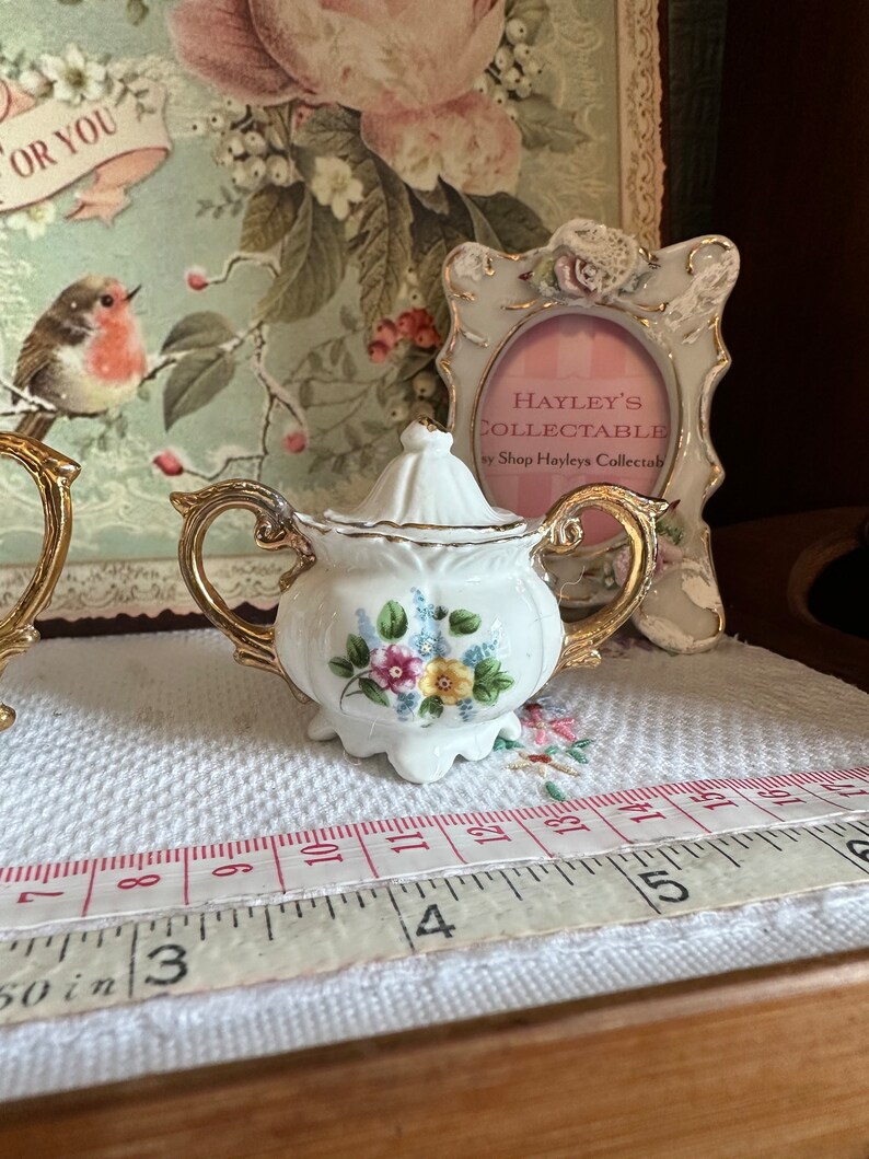 May include: A small, white porcelain sugar bowl with a lid, gold trim, and floral accents. The sugar bowl is approximately 3 inches wide. The background includes a decorative frame and a vintage-style print.