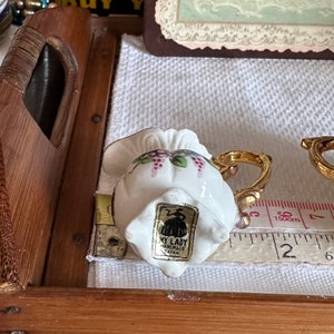 May include: A small, white ceramic teacup with a gold handle and floral design, including purple grapes. The teacup has a label that reads "MY LADY HANDMADE JAPAN". A measuring tape shows the cup is approximately 2 inches wide.