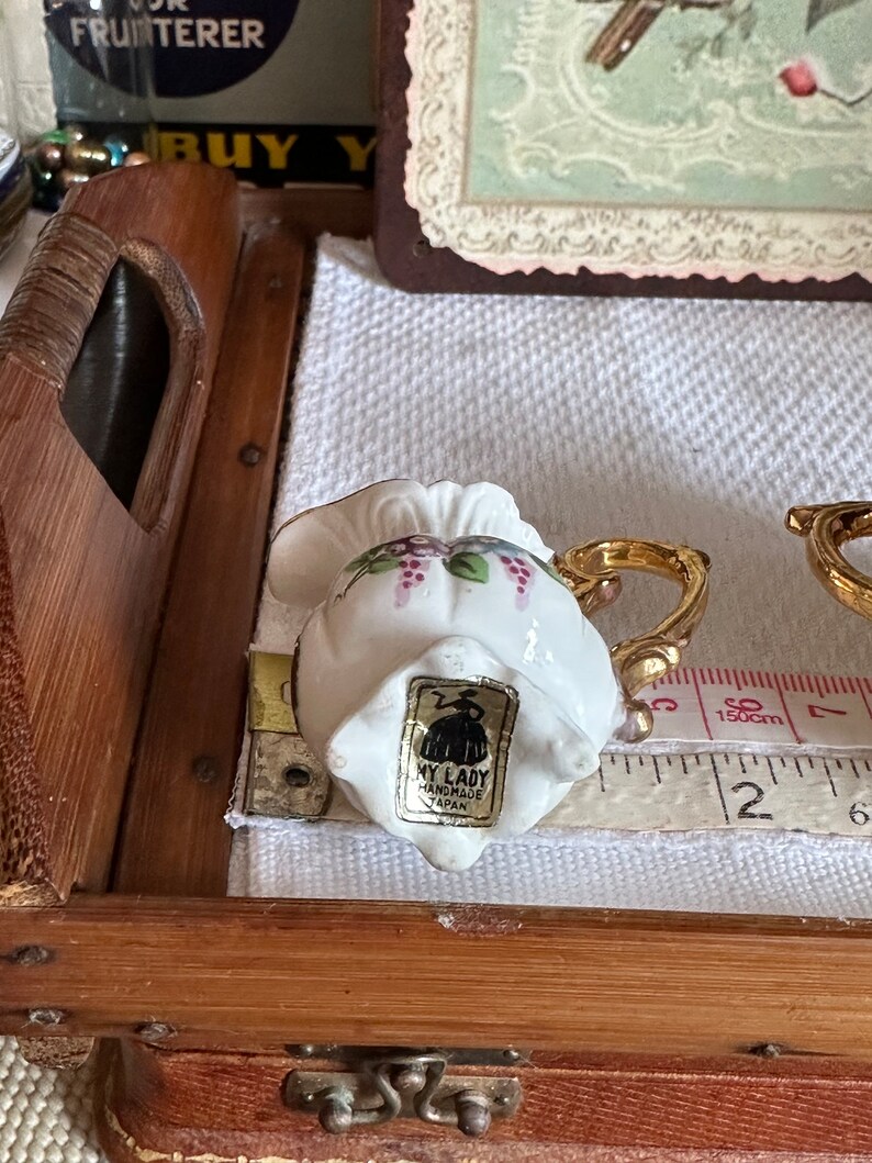 May include: A small, white ceramic teacup with a gold handle and floral design, including purple grapes. The teacup has a label that reads "MY LADY HANDMADE JAPAN". A measuring tape shows the cup is approximately 2 inches wide.