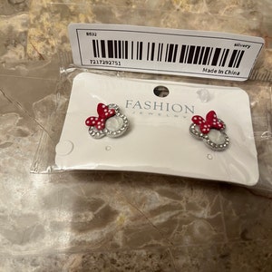 May include: A pair of silver earrings with red and white Minnie Mouse heads. The earrings are on a white card with the words "FASHION JEWELRY" printed on it. The card also has a barcode and the text "Made in China".