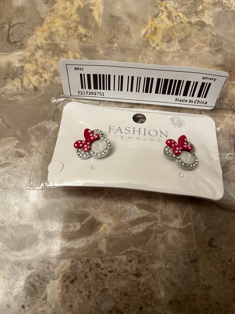 May include: A pair of silver earrings with red and white Minnie Mouse heads. The earrings are on a white card with the words "FASHION JEWELRY" printed on it. The card also has a barcode and the text "Made in China".