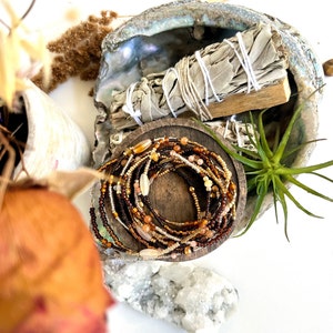 May include: A close-up of a brown, beige, and gold beaded necklace wrapped around a wooden spool. The necklace is on a white surface with a seashell and a bundle of sage.