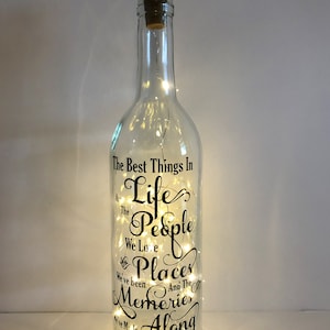 May include: A clear glass bottle with a cork top and a string of lights inside. The bottle has a black vinyl decal with the text "The Best Things In Life Are The People We Love The Places We've Been And The Memories We've Made Along The Way".