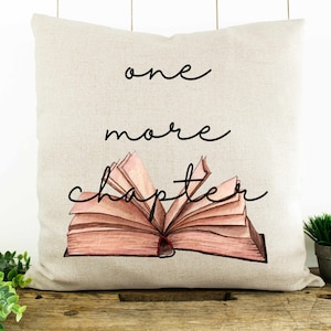 May include: A beige pillow with the text "one more chapter" printed in black cursive. An illustration of an open book with pink pages is centered on the pillow.