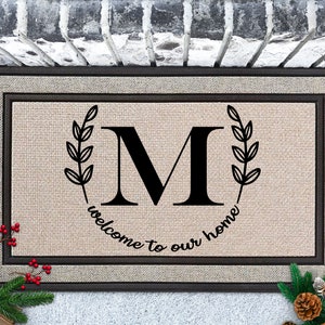 May include: A brown doormat with a black border and a white background. The doormat has a black letter "M" surrounded by two black wreaths with leaves. The text "welcome to our home" is written in a black cursive font around the letter "M".