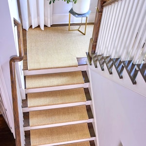 Peut inclure: Un ensemble de tapis d'escalier rectangulaires en tissage beige install&eacute;s sur un escalier en bois avec des contremarches blanches. Les tapis pr&eacute;sentent un aspect textur&eacute; en fibre naturelle et couvrent le centre de chaque marche.