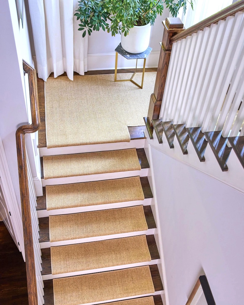 Peut inclure: Un ensemble de tapis d'escalier rectangulaires en tissage beige install&eacute;s sur un escalier en bois avec des contremarches blanches. Les tapis pr&eacute;sentent un aspect textur&eacute; en fibre naturelle et couvrent le centre de chaque marche.