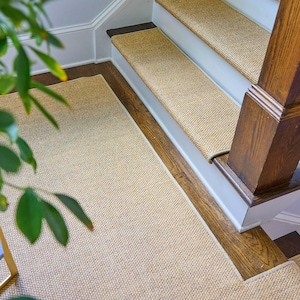 Peut inclure: Un ensemble de tapis d'escalier tiss&eacute;s couleur beige et un tapis de sol rectangulaire assorti. Les tapis d'escalier sont install&eacute;s sur des marches en bois avec des contremarches blanches, tandis que le tapis de sol est pos&eacute; sur le palier.