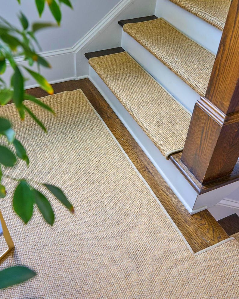 Peut inclure: Un ensemble de tapis d'escalier tiss&eacute;s couleur beige et un tapis de sol rectangulaire assorti. Les tapis d'escalier sont install&eacute;s sur des marches en bois avec des contremarches blanches, tandis que le tapis de sol est pos&eacute; sur le palier.