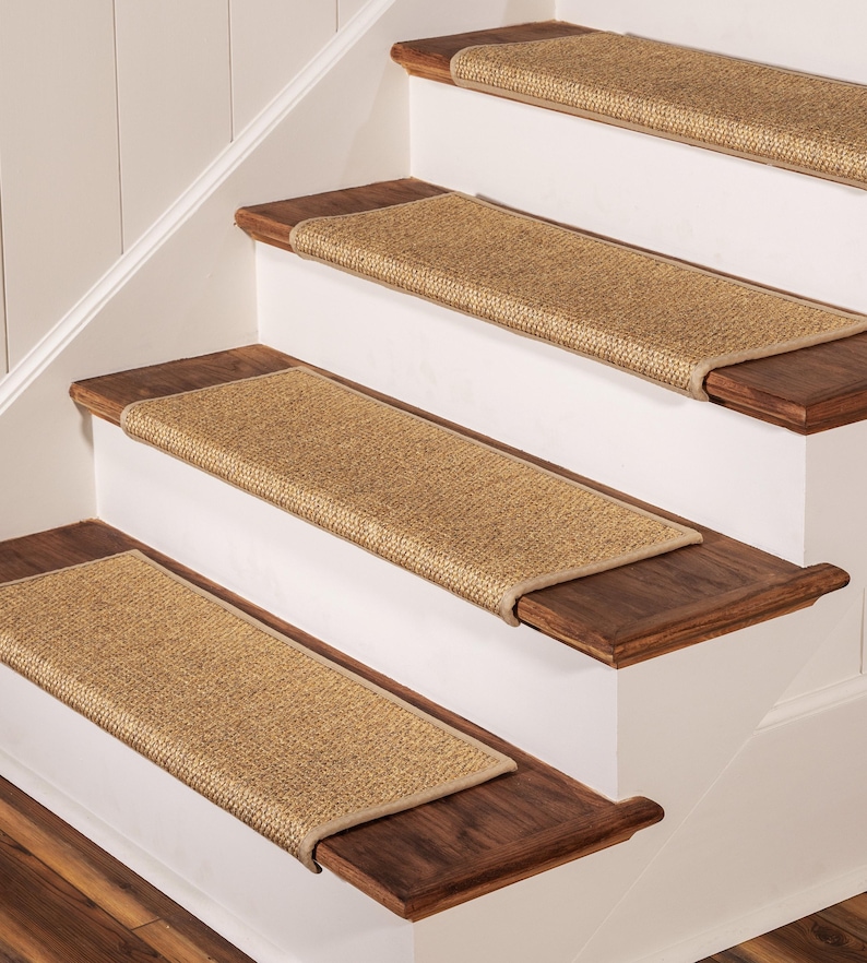Peut inclure: Quatre tapis d'escalier rectangulaires fabriqu&eacute;s dans une mati&egrave;re en fibre naturelle tiss&eacute;e de couleur beige. Chaque tapis pr&eacute;sente un bord fini et gans&eacute; d'une couleur beige assortie, pos&eacute; &agrave; plat au centre de marches en bois brun fonc&eacute;.