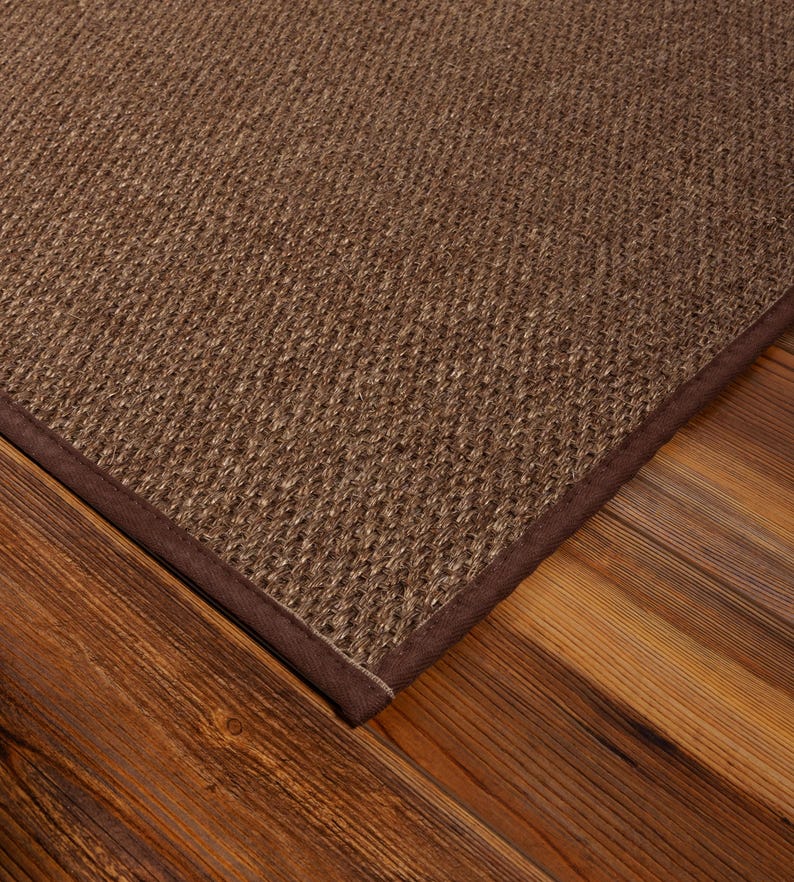 Peut inclure: Un tapis rectangulaire pr&eacute;sentant une surface tiss&eacute;e brune textur&eacute;e avec un motif en grille serr&eacute; et uniforme. Le tapis est fini avec une bordure en tissu marron fonc&eacute;, lisse et solide, qui longe le bord, offrant un cadre net et d&eacute;fini.