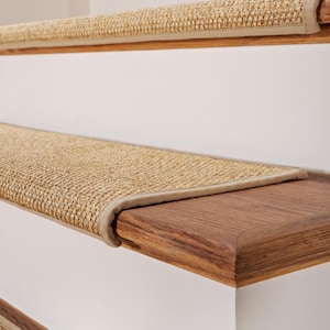 Peut inclure: Un ensemble de tapis d'escalier tiss&eacute;s de couleur beige avec un aspect textur&eacute; en fibres naturelles. Chaque tapis pr&eacute;sente un bord fini avec une bordure en tissu beige assortie, con&ccedil;u pour couvrir la surface horizontale des marches en bois.