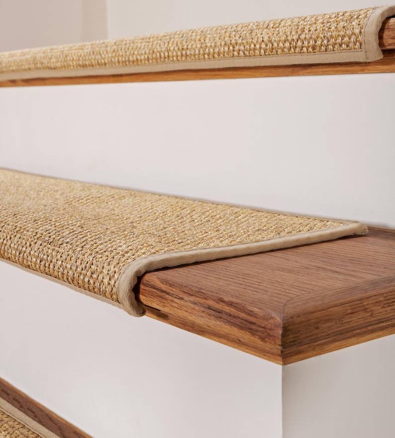 Peut inclure: Un ensemble de tapis d'escalier tiss&eacute;s de couleur beige avec un aspect textur&eacute; en fibres naturelles. Chaque tapis pr&eacute;sente un bord fini avec une bordure en tissu beige assortie, con&ccedil;u pour couvrir la surface horizontale des marches en bois.