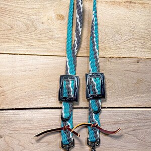 May include: A pair of turquoise, gray, and white braided leather reins with silver buckles and a brown leather tie. The reins have a snap hook on each end.