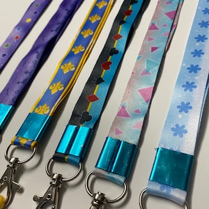 Pu&ograve; includere: Cinque cordini in tessuto con morsetti blu metallizzato e moschettoni girevoli argentati. I motivi includono viola con stelle, giallo con gigli, nero con semi di carte, azzurro con triangoli geometrici e azzurro con forme di fiori.