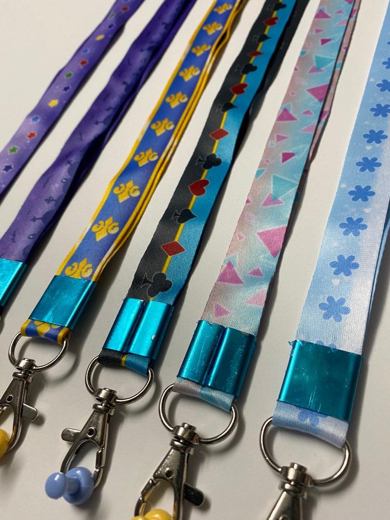 Pu&ograve; includere: Cinque cordini in tessuto con morsetti blu metallizzato e moschettoni girevoli argentati. I motivi includono viola con stelle, giallo con gigli, nero con semi di carte, azzurro con triangoli geometrici e azzurro con forme di fiori.