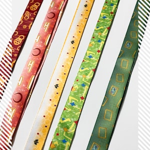 Pu&ograve; includere: Cinque lunghi cordini in tessuto con motivi distinti: una stampa con ingranaggi rosso scuro, un design con mezzaluna rosa, un motivo a nido d'ape giallo con piccole api nere, un motivo geometrico verde con stelle colorate e una stampa con cornici rettangolari verde scuro.
