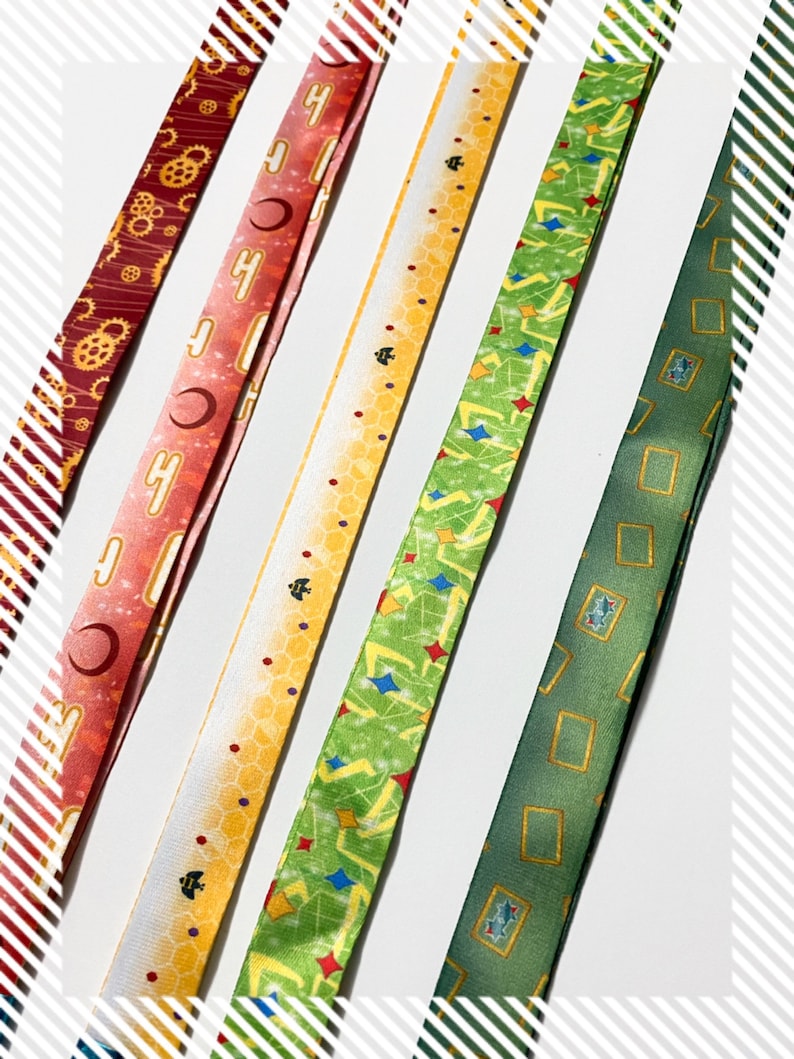 Pu&ograve; includere: Cinque lunghi cordini in tessuto con motivi distinti: una stampa con ingranaggi rosso scuro, un design con mezzaluna rosa, un motivo a nido d'ape giallo con piccole api nere, un motivo geometrico verde con stelle colorate e una stampa con cornici rettangolari verde scuro.