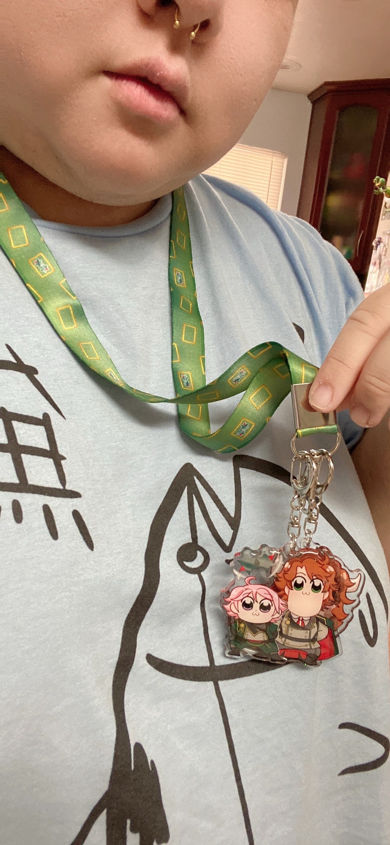 Pu&ograve; includere: Un cordino verde con un motivo rettangolare giallo ripetuto attaccato a un portachiavi in acrilico trasparente raffigurante due personaggi dei cartoni animati con capelli rosa e arancioni, che indossano uniformi militari, sospeso da una catena in metallo argentato.