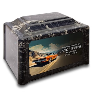 May include: A black marble cremation urn with a rectangular shape and a decorative top. The urn features a color image of an orange car on a road with mountains in the background. Text reads "IN LOVING MEMORY OF JACK STEVENS WE MISS YOU 11/12/65-12/11/24."