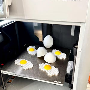 May include: A white 3D printer with a black interior. The printer is open and the build plate is visible. There are five white 3D printed objects on the build plate, including four eggs and one fried egg. The printer has a label that reads "Bambu Lab X1-Carbon".
